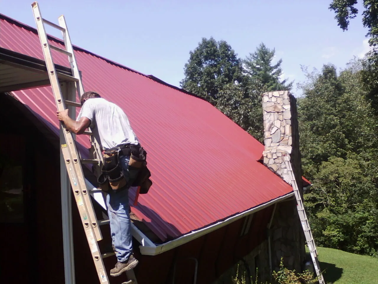 Roofer on a ladder beside a red metal roof and stone chimney — Soffit & Fascia Repair by Roofing Brothers in Great Neck